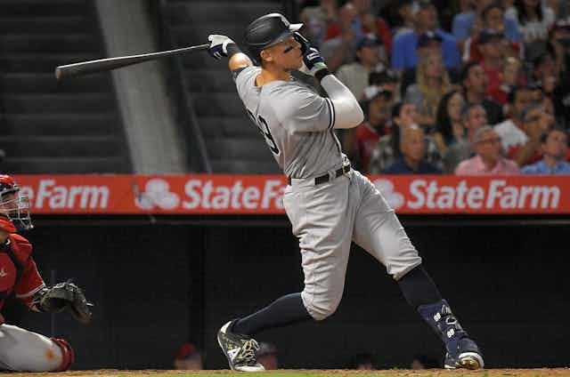A baseball player in a Yankees jersey looks toward the field as he finishes his swing. The crowd behind him is looking in the same direction.