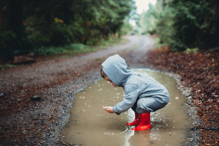 a toddler in boots squats in a puddle on a dirt road in the woods on a rainy day