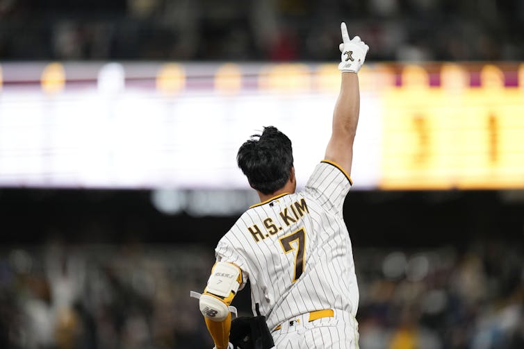 A ball player raises one finger in the air as he runs the bases, with bright stadium lights behind him.