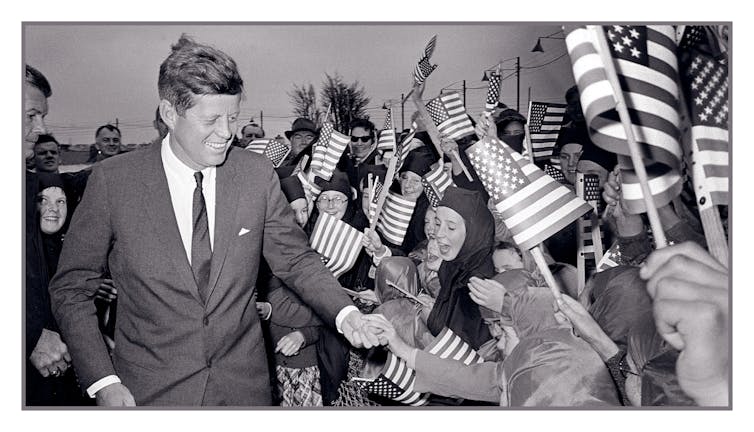 Black and white photo of a smiling President Kennedy greeted by a crowd of cheering children and nuns, all waving small US flags.