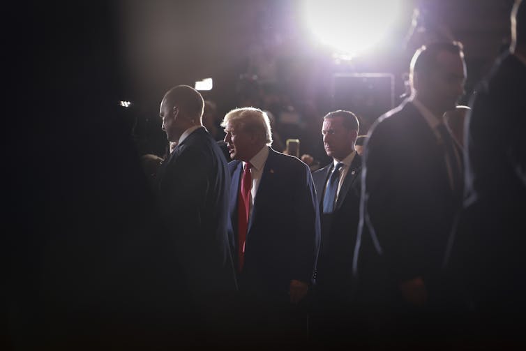 A man in a dark blue suit is seen lit up with artificial lights as he walks surrounded by several men.