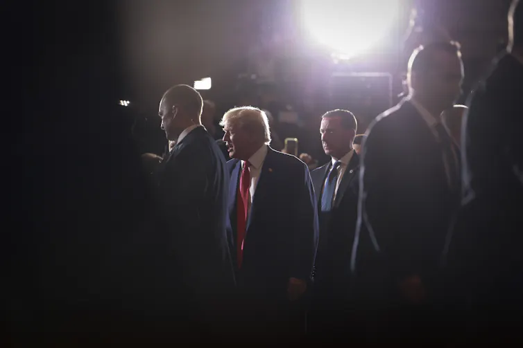 A man in a dark blue suit is seen lit up with artificial lights as he walks surrounded by several men.