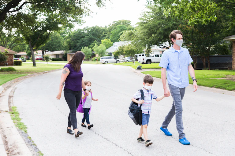 A man and a woman walk a boy and a girl, respectively, home from school. The children are carrying backpacks and holding the adults' hands.