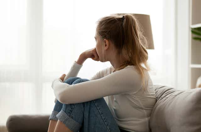 A teen girls sits on a couch looking out a window.