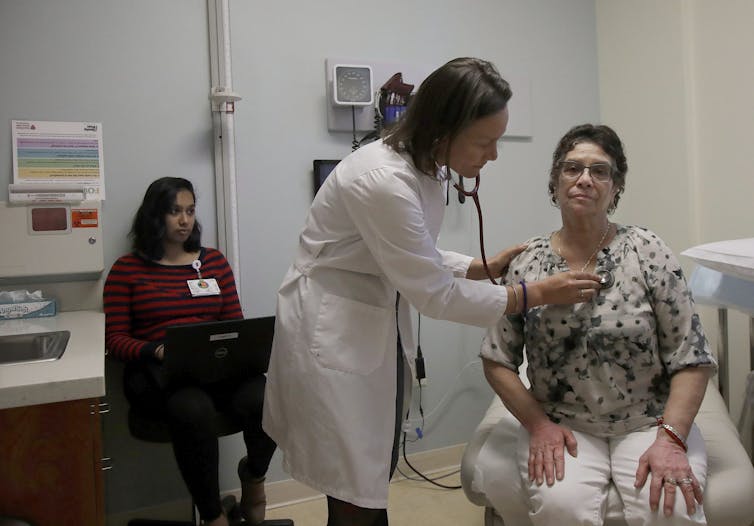 A doctor examining a patient, while a woman typing on a laptop sits in the background