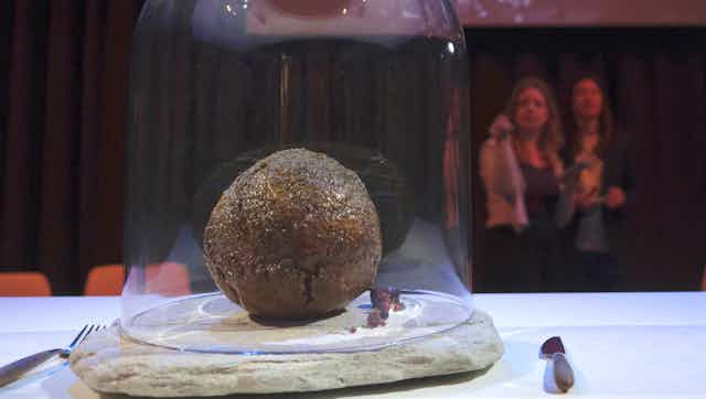 A photo of a meatball made from cultured mammoth meat on a plate beneath a glass cloche with spectators in the background.