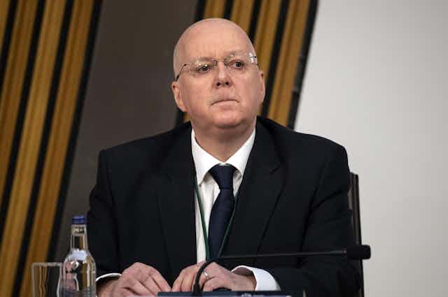 A bald man in a dark suit sitting at a conference table looking sombre.