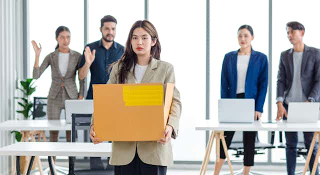 Sad woman in casual suit standing holding belongings in cardboard box after fired while male and female colleagues wave goodbye in blurred background.