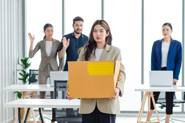 Sad woman in casual suit standing holding belongings in cardboard box after fired while male and female colleagues wave goodbye in blurred background.