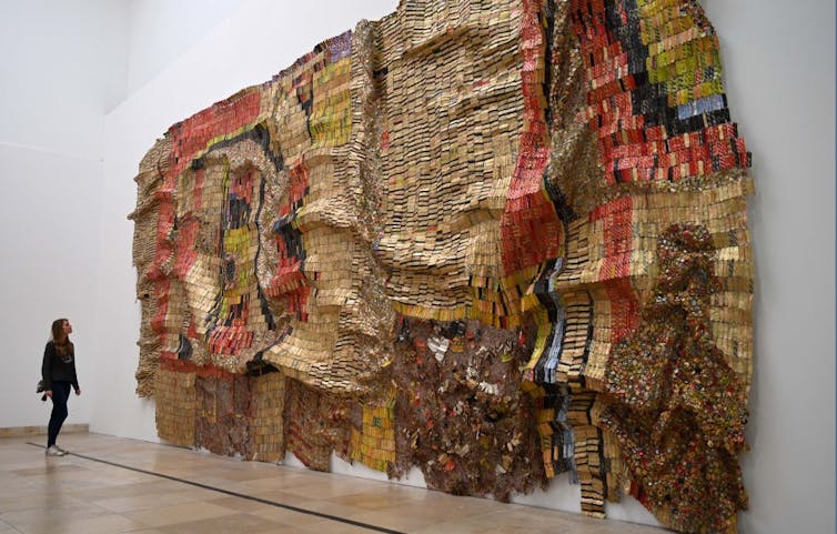 A person is dwarfed by the enormous scale of an artwork on a wall, glimmering in coppers, bronzes and golds, a folded hanging made of thousands of different types of metal.
