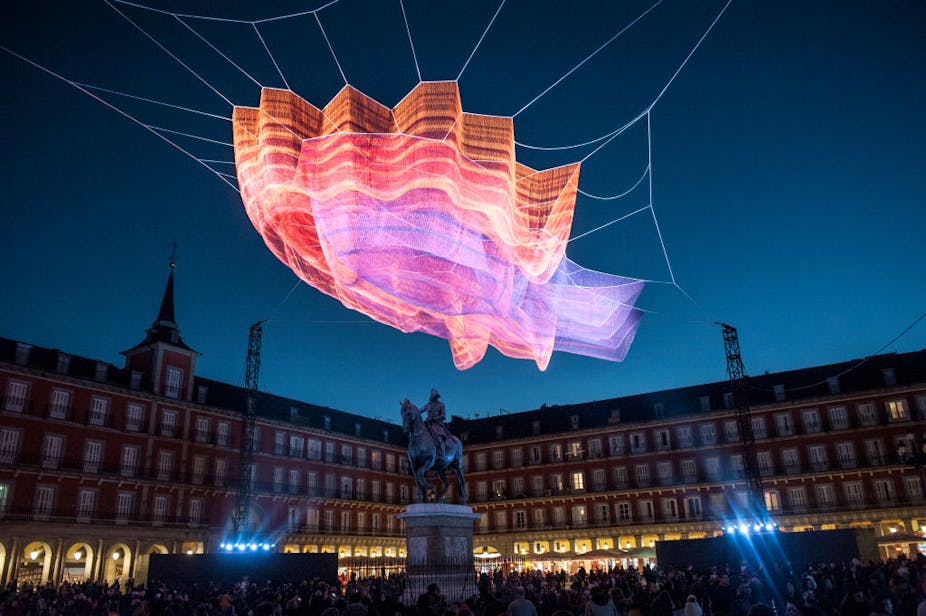 Large pink, red and orange sculpture hangs above buildings and a crowd of people at night.