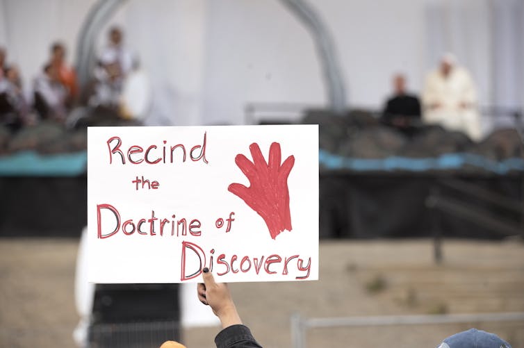 A protest sign is held up. It says: Rescind the Doctrine of Discovery (sic).