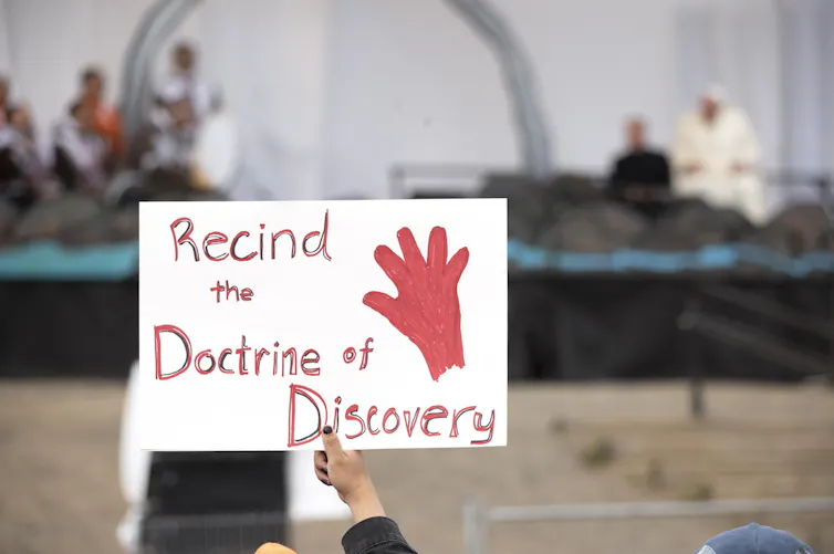 A protest sign is held up. It says: Rescind the Doctrine of Discovery (sic).