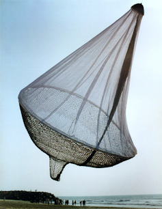 Large sculpture made of netting hangs above beach.