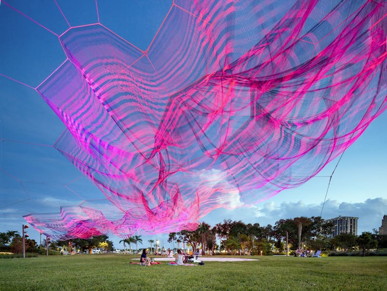 Large pink and purple sculpture suspended over a green field with blue sky in the background.