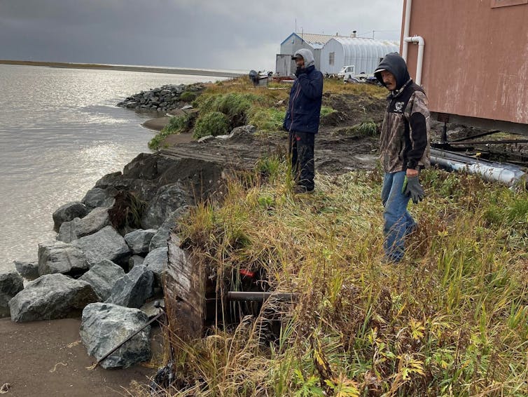 Two men look at erosion that has exposed several feet of pipe. Rocks help protect part of the property.