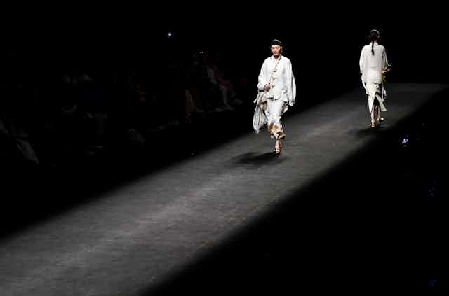 Two women dressed in white walk along the runway during a fashion show.