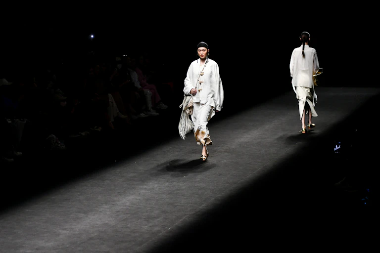 Two women dressed in white walk along the runway during a fashion show.