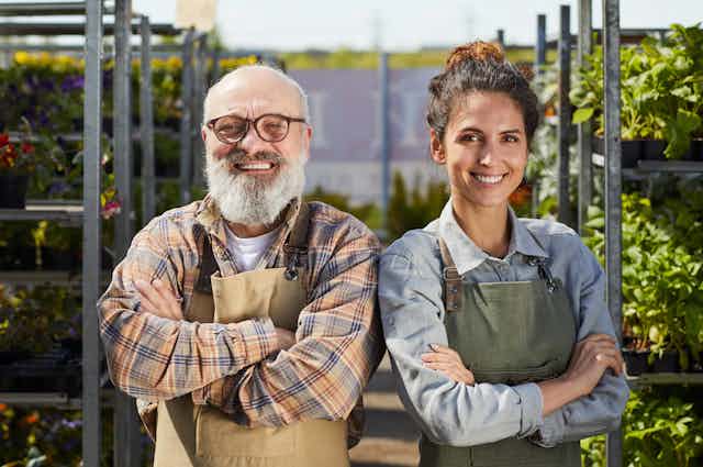 A bald man with a white beard and glasses stands next to a young woman with her hair tied back. Both have their arms crossed and are wearing aprons. They are standing in front of rows of potted plants.