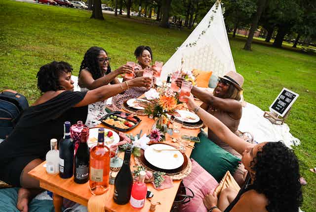 A group of Black women friends, raise their drinks in cheers as they enjoy a meal at a picnic table.