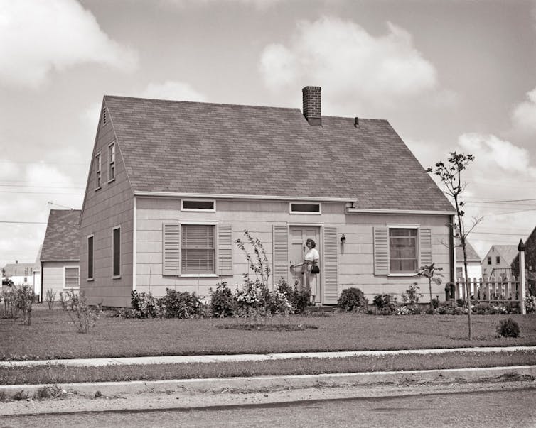Black and white photo of woman standing outside her suburban home with a perfectly manicured lawn.
