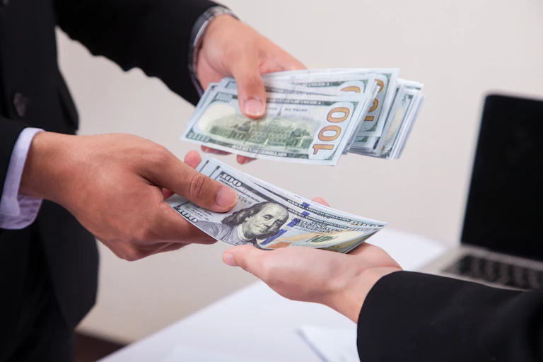 closeup shows someone hands as he puts $100 bills in someone else's hands one by one