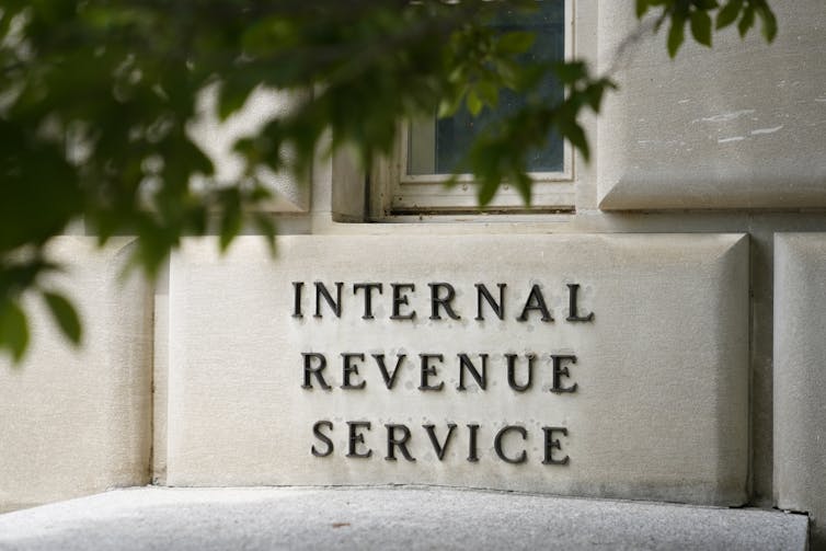 a stone block has the letters internal revenue service carved into it behind the green leaves of a tree