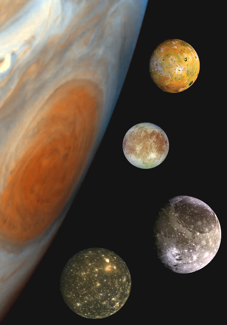 Four moons next to a large red spot on the surface of Jupiter.