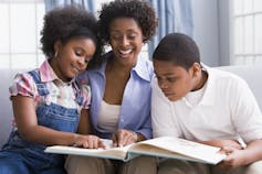 A Black mother looks at a book along with her two children, a girl and a boy, while seated on a couch in their home.