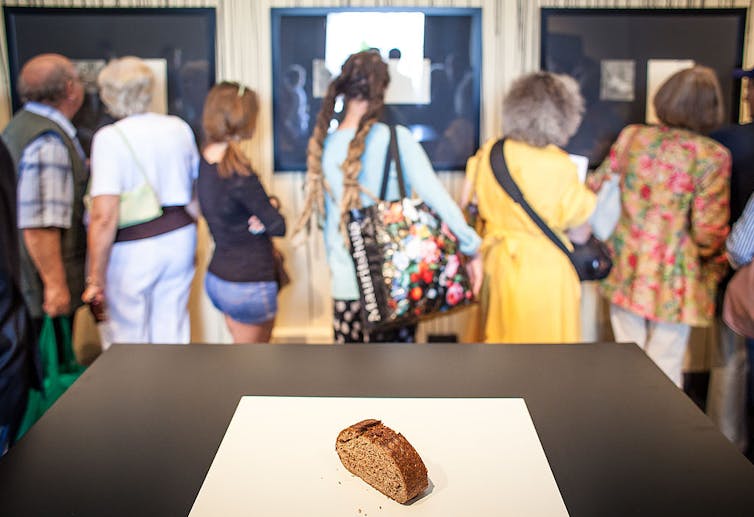 People look at a museum display. In the foreground, a single slice of bread sits on a table.