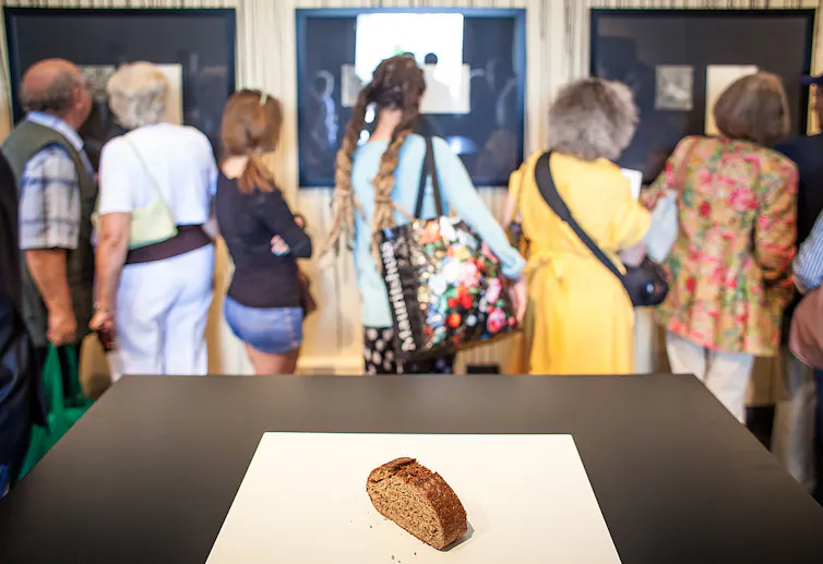 People look at a museum display. In the foreground, a single slice of bread sits on a table.