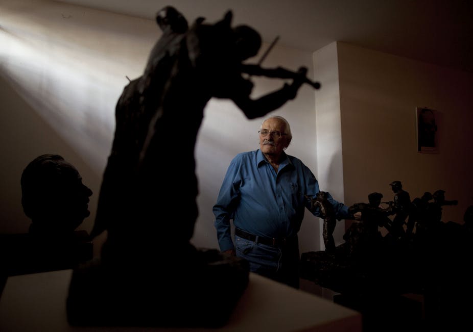 An older man with glasses and a blue collared shirt stands in the back of a room with fading sunlight and small sculptures.