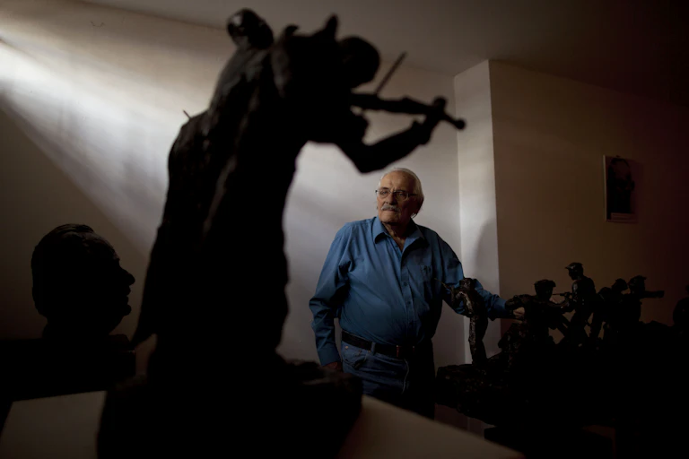 An older man with glasses and a blue collared shirt stands in the back of a room with fading sunlight and small sculptures.