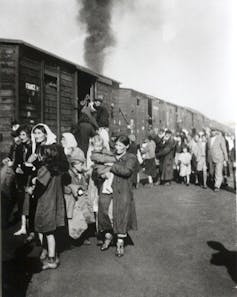 A black and white photo shows women and children in coats walking beside cattle cars.