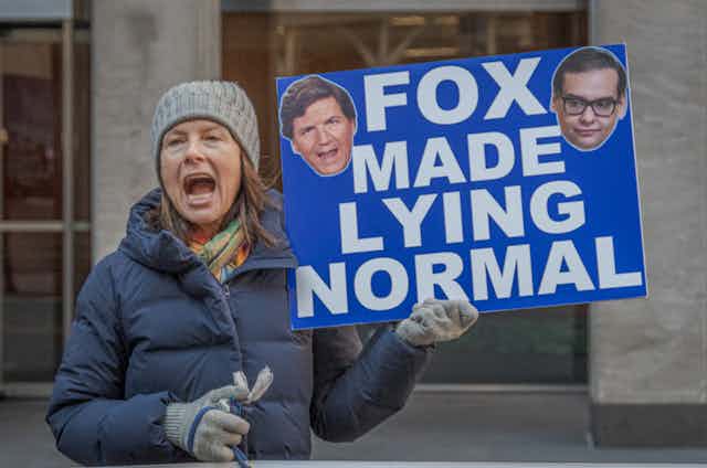 A woman dressed for winter holding a sign that says 'Fox made lying normal.'