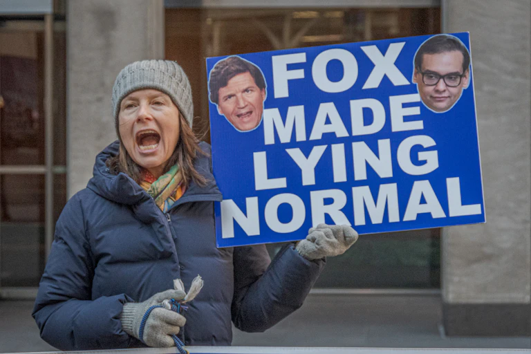 A woman dressed for winter holding a sign that says 'Fox made lying normal.'