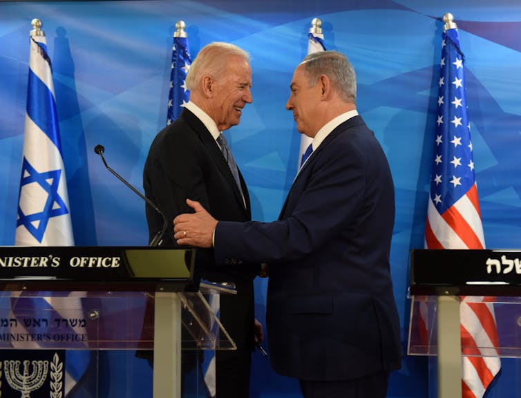 Two older men shake hands and smile, standing in front of a blue backdrop and American and Israeli flags.