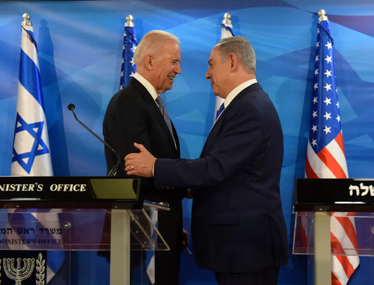 Two older men shake hands and smile, standing in front of a blue backdrop and American and Israeli flags.