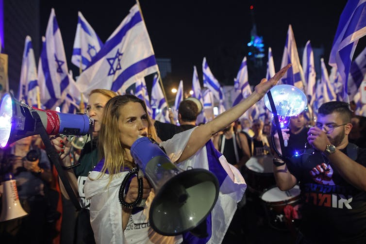 Una mujer joven parece gritar por un megáfono y levanta el brazo, rodeada de otras personas, todas con banderas israelíes.