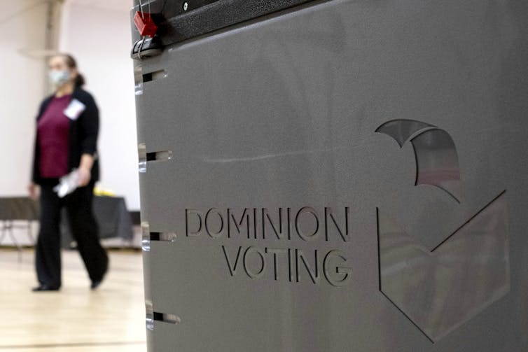 A machine with the words'Dominion Voting' on it, and a woman walking by in the background.