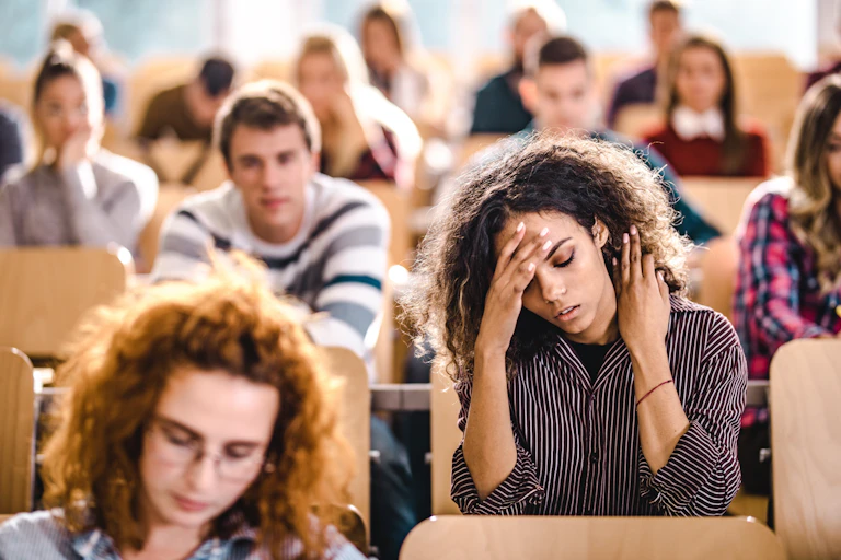 A female college student rubs her temple with one hand and neck with the other as her eyes are closed. She sits among other students in a lecture hall.