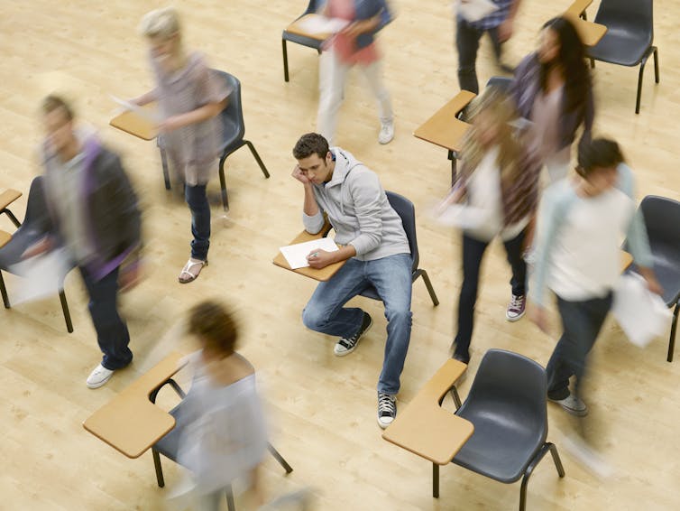 A distressed-looking male college student sits at a desk in a blur of other students.