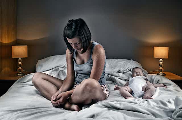 A woman sits on her bed cross-legged, looking downcast with her back turned to her infant.