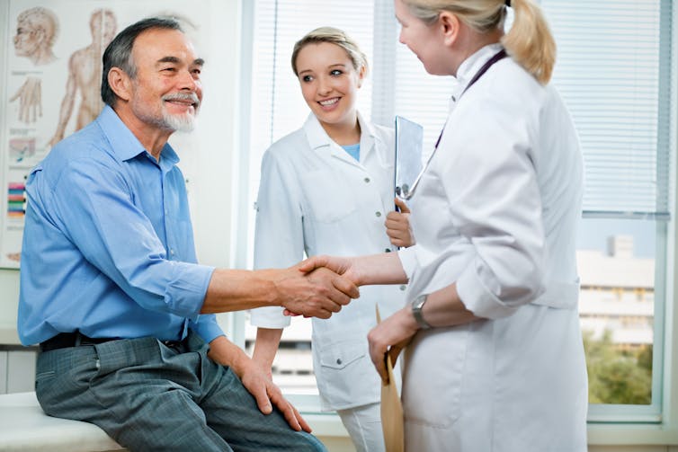 A doctor shakes a patient's hand while a young woman with a clipboard watches