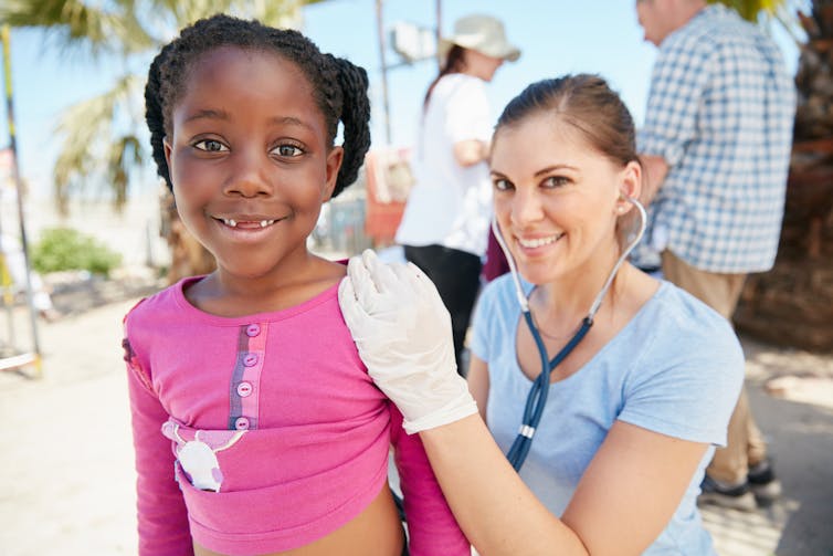 A child in the foreground as a young woman wearing a stethoscope smiles behind her