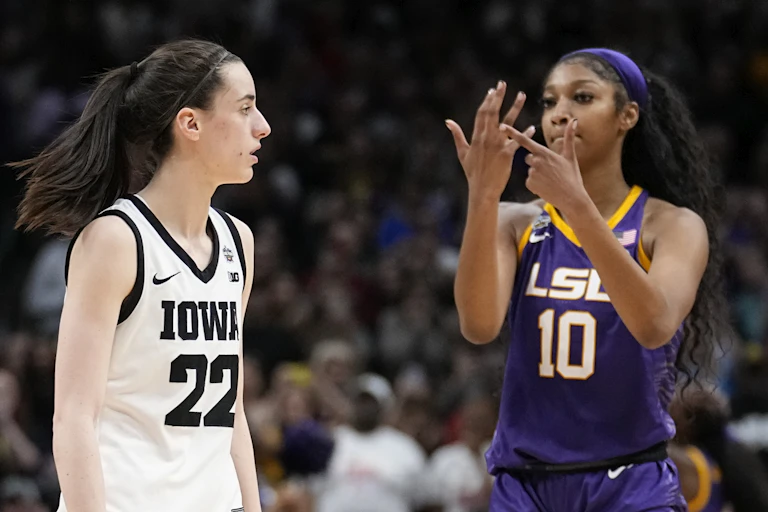A woman in a basketball uniform makes a hand gesture as another player walks past.