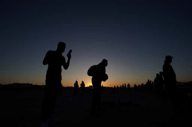 People are seen in silhouette against a sunset. One looks at his phone.