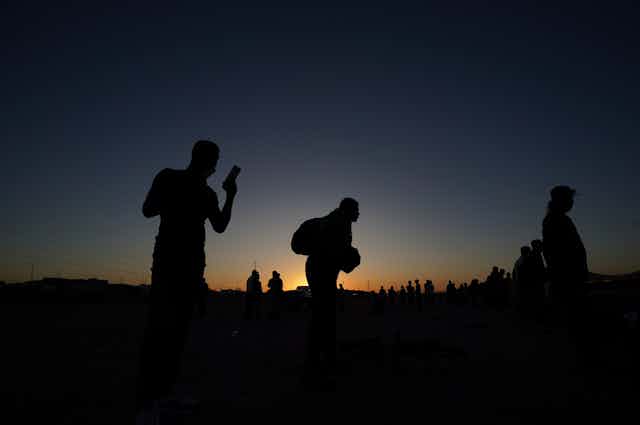 People are seen in silhouette against a sunset. One looks at his phone.