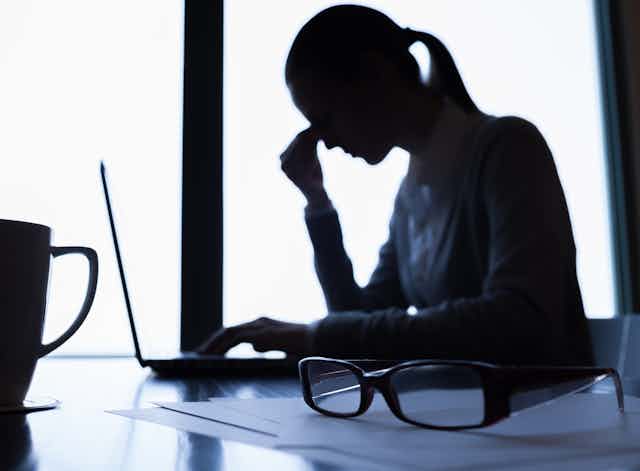 The silhouette of a woman pinching the bridge of her nose while sitting in front of an open laptop