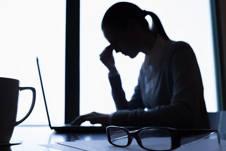 The silhouette of a woman pinching the bridge of her nose while sitting in front of an open laptop